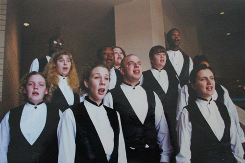 Students in white dress shirts and black vests singing together in a choir recital.