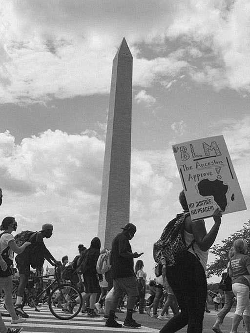 Black and white photograph of march participants; the Washington Monument looms in the background. One participant in the foreground holds a poster with an image of Africa that says BLM The ancestors approve: no justice, no peace.