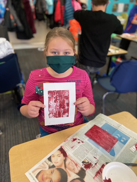 Child in a face mask holds up a red print that reads Kindness.