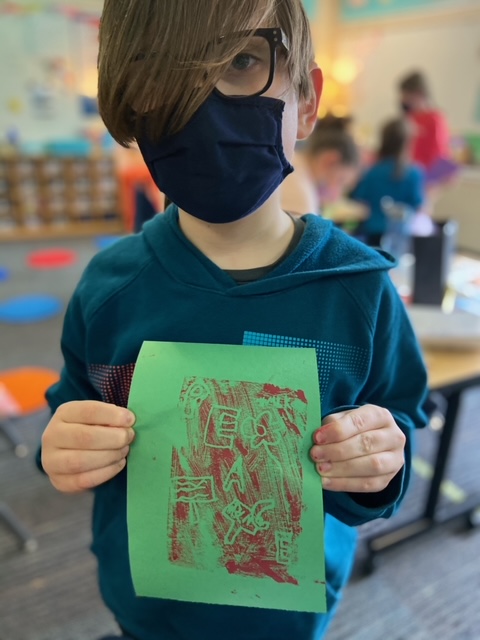 Child in a face mask holds up a red print on green paper that reads, Peace.