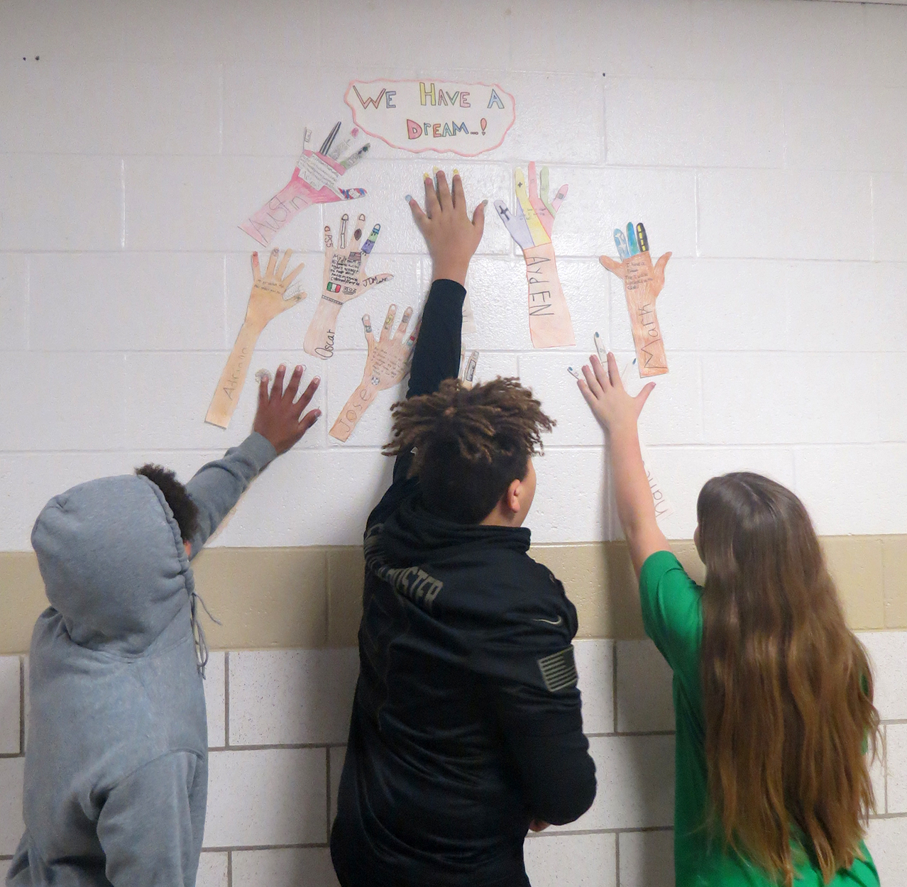 Wall of art - 3 students reach up to put their hands next to tracings of hands with drawings inside.