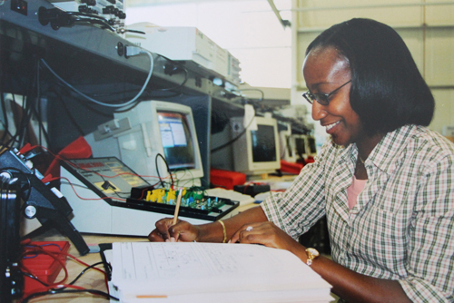 A student writes in a notebook surrounded by computers and equipment.