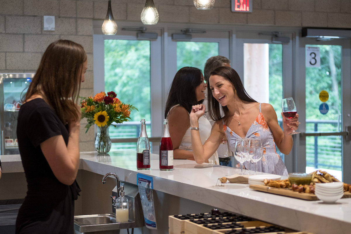Three women stand at a modern indoor bar counter, chatting and smiling. One woman in a floral dress holds a glass of red wine and gestures animatedly while talking. Bottles of rosé, empty wine glasses, and a charcuterie board with snacks sit on the white countertop. A vase of colorful flowers decorates the space, and glass doors in the background reveal greenery outside. Warm pendant lights hang overhead, creating a relaxed, social atmosphere.