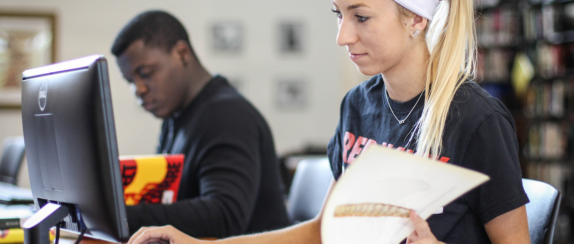 Two students are studying with their books in front of computers. 