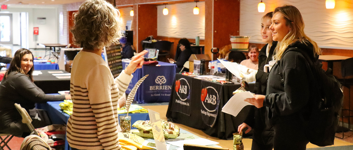 A group of people interact at a job or resource fair. A woman with curly hair stands at a booth covered with pamphlets, shirts, and display materials, speaking with two young women holding papers. Other booths with banners and seated representatives are visible in the background inside a well-lit indoor space.