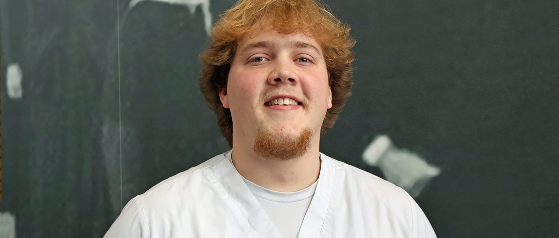 Cameron Schinck wearing white medical scrubs smiles at the camera while standing in front of a dark chalkboard background with faint chalk marks.