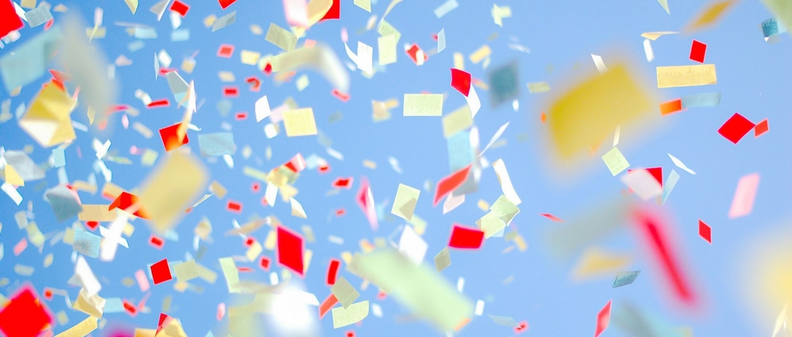 Colorful paper confetti in red, yellow, and white floats through the air against a bright blue sky, creating a festive, celebratory scene.
