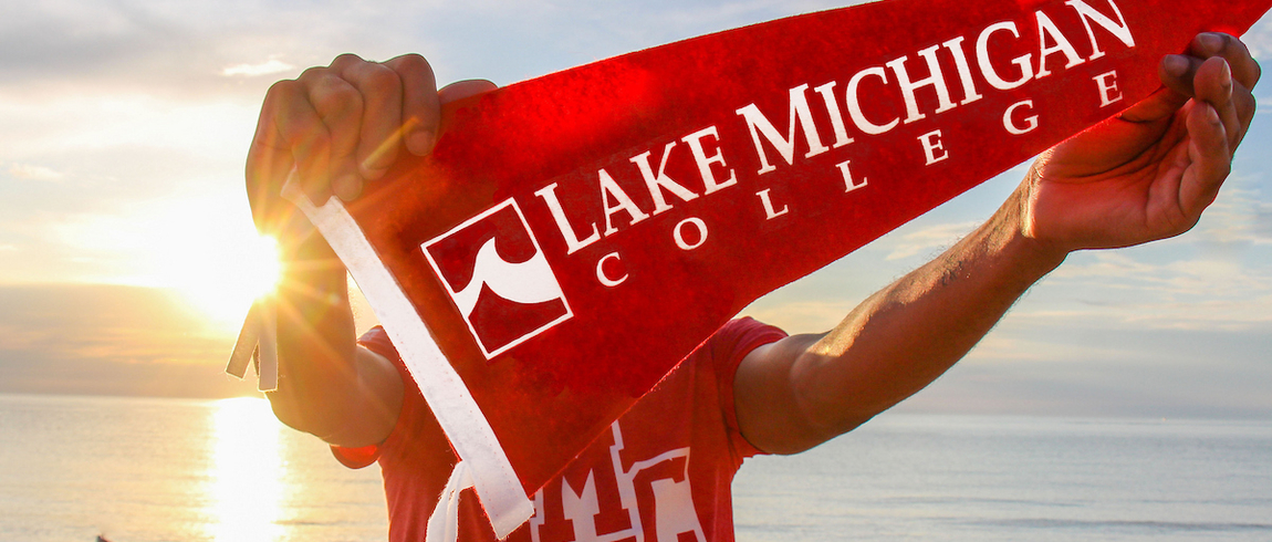 A person standing on a sandy beach at sunset holds a red Lake Michigan College pennant toward the camera, with the sun glowing brightly behind them over the water.