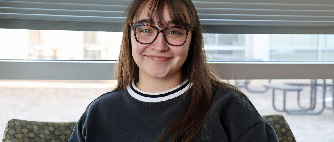 A smiling young woman with long brown hair and glasses sits indoors in front of a window, wearing a dark crewneck sweatshirt.