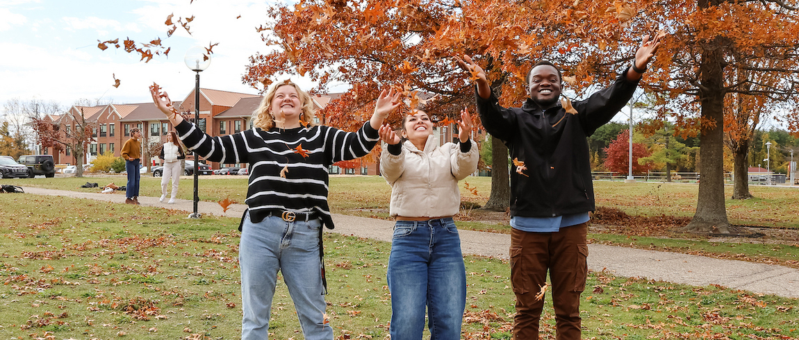 Three students stand on a grassy campus lawn in autumn, smiling and throwing orange leaves into the air beneath a large tree with fall foliage, with academic buildings and a walkway in the background.