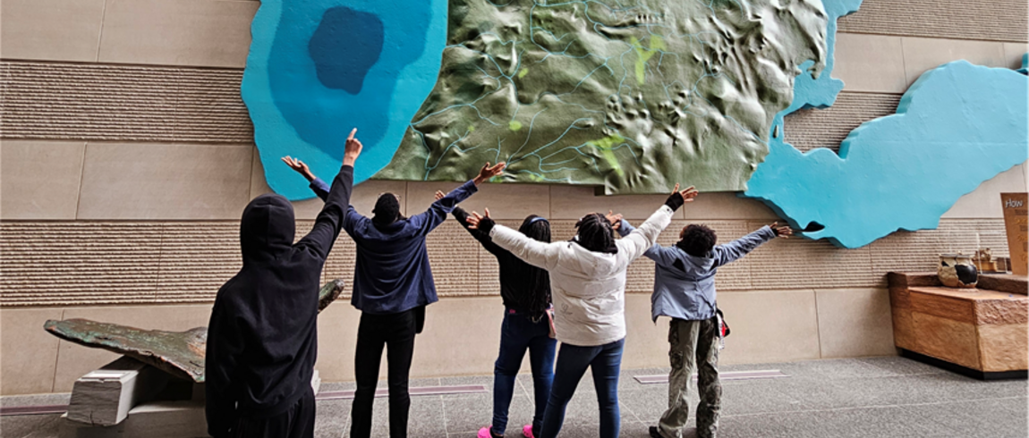 Five students stand facing a large, wall-mounted 3D relief map of Michigan and the surrounding Great Lakes. With their backs to the camera, they raise their arms toward the map as if pointing or celebrating. The map shows detailed terrain in green and blue water areas.