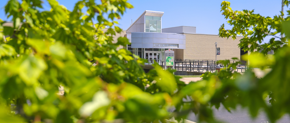 Entrance of Lake Michigan College campus building on a sunny day, viewed through green leaves in the foreground.
