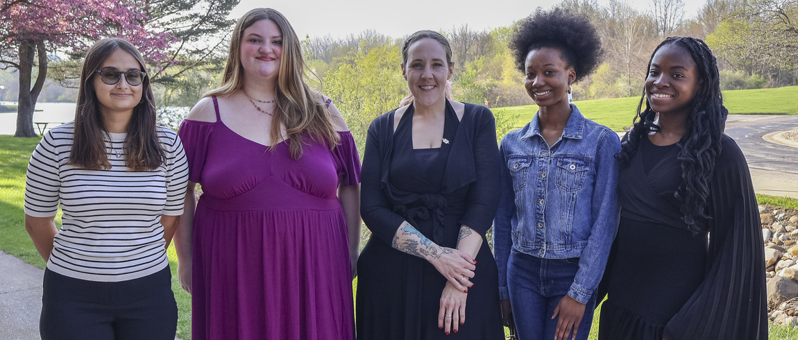 Five women stand side by side outdoors on a sunny day, smiling at the camera.