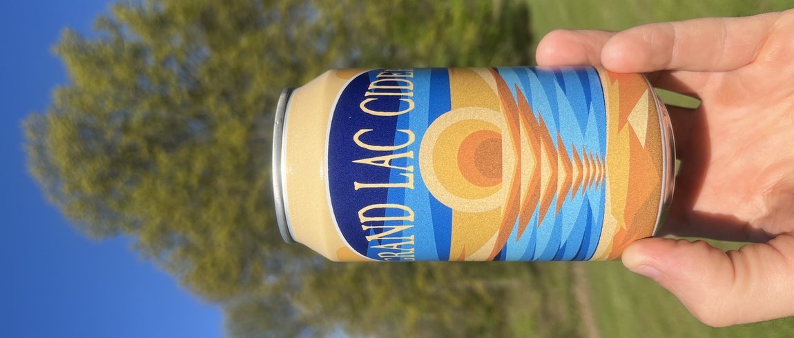 A can of Grand Lac Cider is held up in front of grass and trees on a sunny day, with a blue sky in the background. 