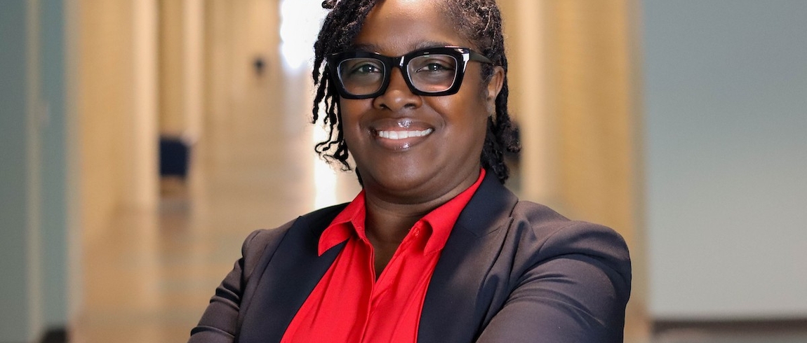 Woman smiles in foreground of picture wearing glasses, red shirt, and suit jacket. Behind her is a long hallway, at the end of which is a window with sunlight shining in. 