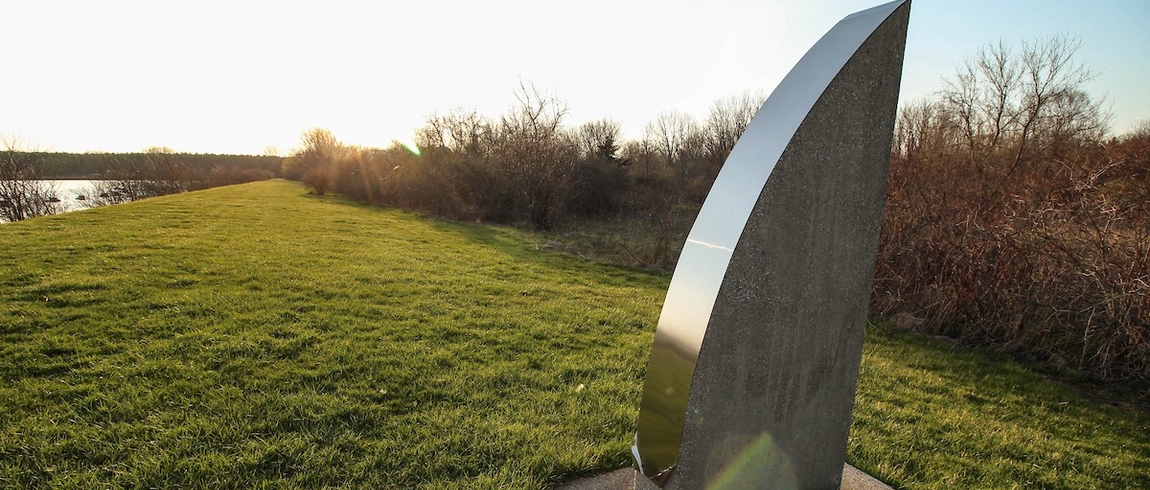 Geometric sculpture on green grass, with a blue sky, trees, and water in the background. 