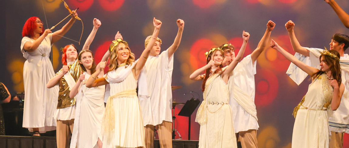 Multiple performers in toga costumes with gold-leaf headbands are posed on stage, each as if aiming a bow and arrow upward. One performer in the back is holding an actual bow-and-arrow prop and appears to be singing.