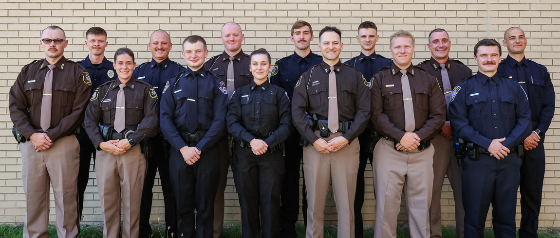 Fourteen men and women in uniform stand on the grass outside in front of a brick wall, looking at the camera. 