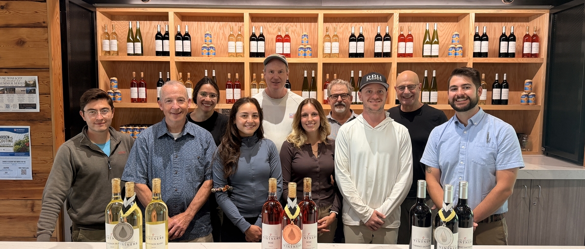 A group of people stands smiling behind a counter with wine bottles draped in three medals. Wine bottles fill open shelves in the background. 