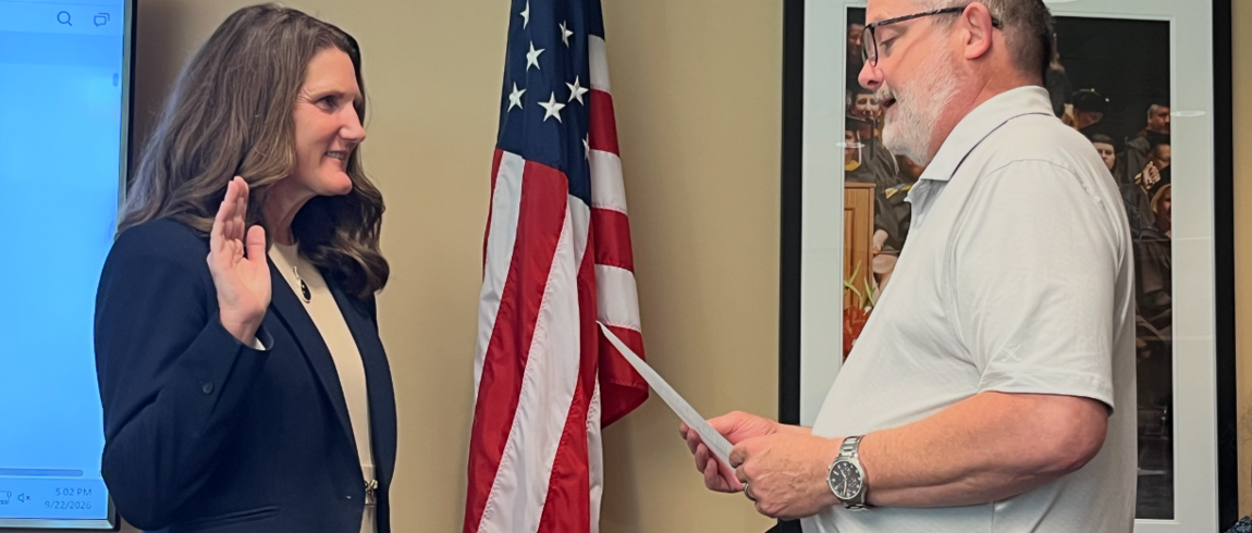 Two individuals standing in front of a flag, one with her hand raised and the other reading from a document.