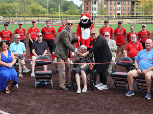 Photo:  Jeremy Miller, Liz Miller (seated), and Jennifer Hoover, surrounded by family, Lake Michigan College officials, and the LMC softball team, prepare to cut the ribbon during the dedication ceremony of the Liz and Lloyd Miller Family Field on the LMC Benton Harbor campus.