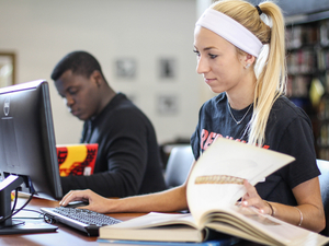 Two students are studying with their books in front of computers. 