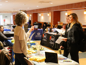 A group of people interact at a job or resource fair. A woman with curly hair stands at a booth covered with pamphlets, shirts, and display materials, speaking with two young women holding papers. Other booths with banners and seated representatives are visible in the background inside a well-lit indoor space.