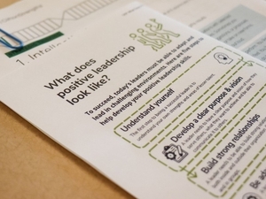 Papers on table. Text in focus reads, "What does positive leadership look like? To succeed, today's leaders must be able to adapt and lead in challenging environments. Here are five steps to help develop your positive leadership skills. Understand yourself. Develop a clear purpose & vision. Build strong relationships. Be adaptable."