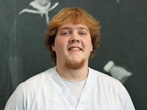 Cameron Schinck wearing white medical scrubs smiles at the camera while standing in front of a dark chalkboard background with faint chalk marks.