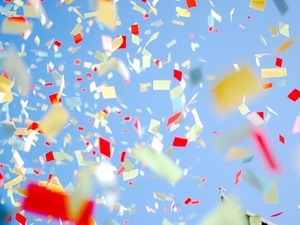 Colorful paper confetti in red, yellow, and white floats through the air against a bright blue sky, creating a festive, celebratory scene.