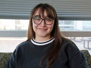 A smiling young woman with long brown hair and glasses sits indoors in front of a window, wearing a dark crewneck sweatshirt.