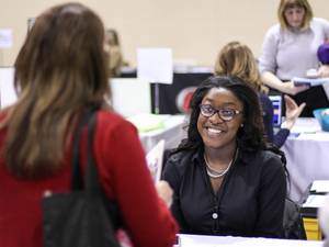 A smiling woman wearing glasses sits behind a table at a job and transfer fair, speaking with another person whose back is facing the camera. The seated woman is dressed in a black blouse and a silver necklace, and she appears engaged and welcoming. In the background, other attendees and staff interact at tables with papers, displays, and informational materials.