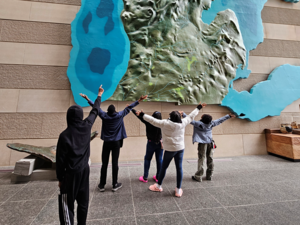 Five students stand facing a large, wall-mounted 3D relief map of Michigan and the surrounding Great Lakes. With their backs to the camera, they raise their arms toward the map as if pointing or celebrating. The map shows detailed terrain in green and blue water areas.
