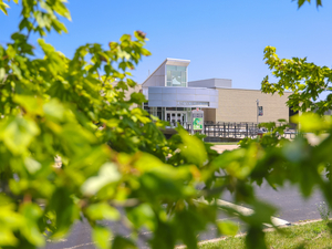 Entrance of Lake Michigan College campus building on a sunny day, viewed through green leaves in the foreground.