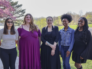 Five women stand side by side outdoors on a sunny day, smiling at the camera.