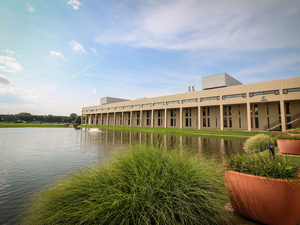Campus building next to water with a blue sky in the background and green plants in the foreground.