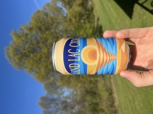 A can of Grand Lac Cider is held up in front of grass and trees on a sunny day, with a blue sky in the background. 