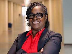 Woman smiles in foreground of picture wearing glasses, red shirt, and suit jacket. Behind her is a long hallway, at the end of which is a window with sunlight shining in. 