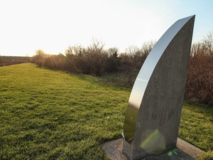 Geometric sculpture on green grass, with a blue sky, trees, and water in the background. 