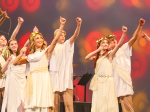 Multiple performers in toga costumes with gold-leaf headbands are posed on stage, each as if aiming a bow and arrow upward. One performer in the back is holding an actual bow-and-arrow prop and appears to be singing.