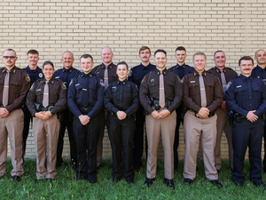 Fourteen men and women in uniform stand on the grass outside in front of a brick wall, looking at the camera. 