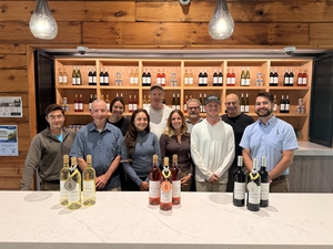A group of people stands smiling behind a counter with wine bottles draped in three medals. Wine bottles fill open shelves in the background. 