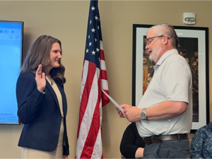 Two individuals standing in front of a flag, one with her hand raised and the other reading from a document.